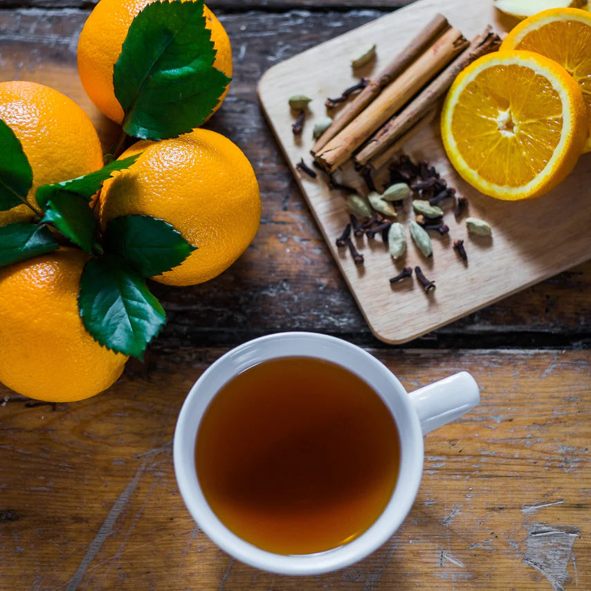 Filiżanka czarnej herbaty z przyprawami, obok pomarańcze, cynamon, kardamon i goździki na drewnianym stole – kompozycja inspirowana herbatą Suki Spiced Citrus.