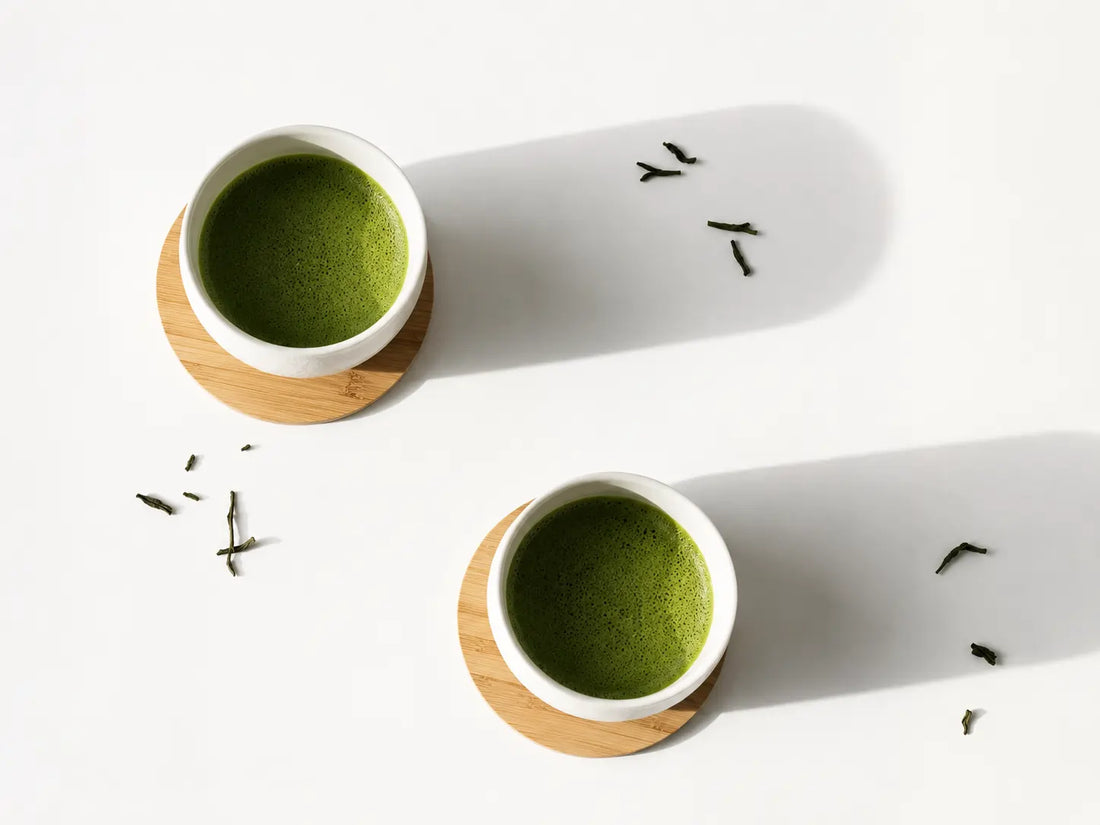 Two bowls of matcha on wooden stands, with scattered tea leaves alongside, set against a white background with a long shadow.