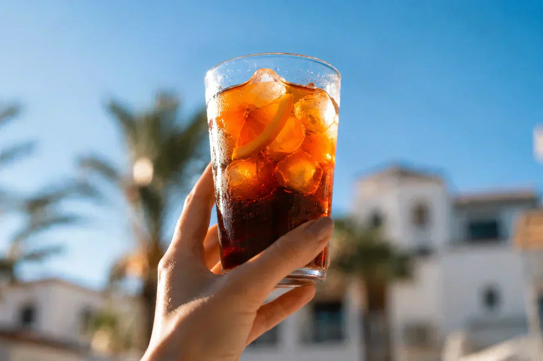 Glass of iced cold brew held in hand against a seaside backdrop with palm trees and white buildings – JAVA Coffee summer mood.