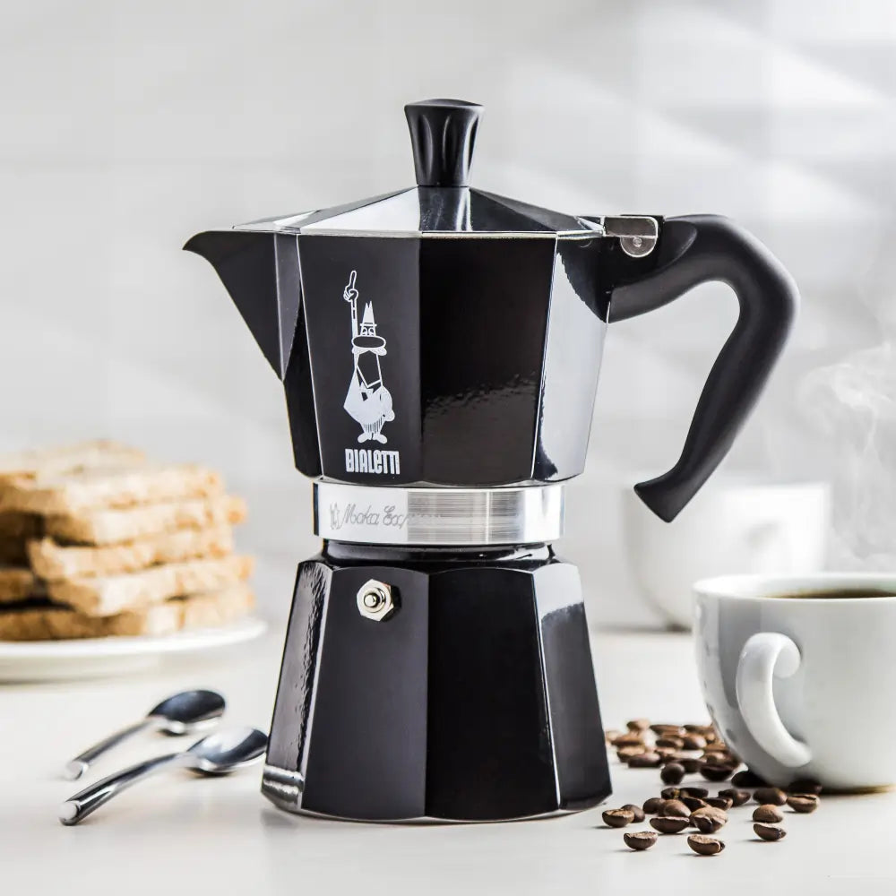  Czarna kawiarka Bialetti Moka Express 6 tz ustawiona na kuchennym blacie obok filiżanki kawy, ziaren i kromek chleba – elegancki design w codziennej aranżacji.