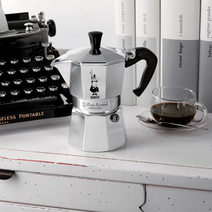 Kawiarka Bialetti Moka Express w stylowej aranżacji retro – ustawiona obok maszyny do pisania, książek i filiżanki czarnej kawy na białej komodzie.