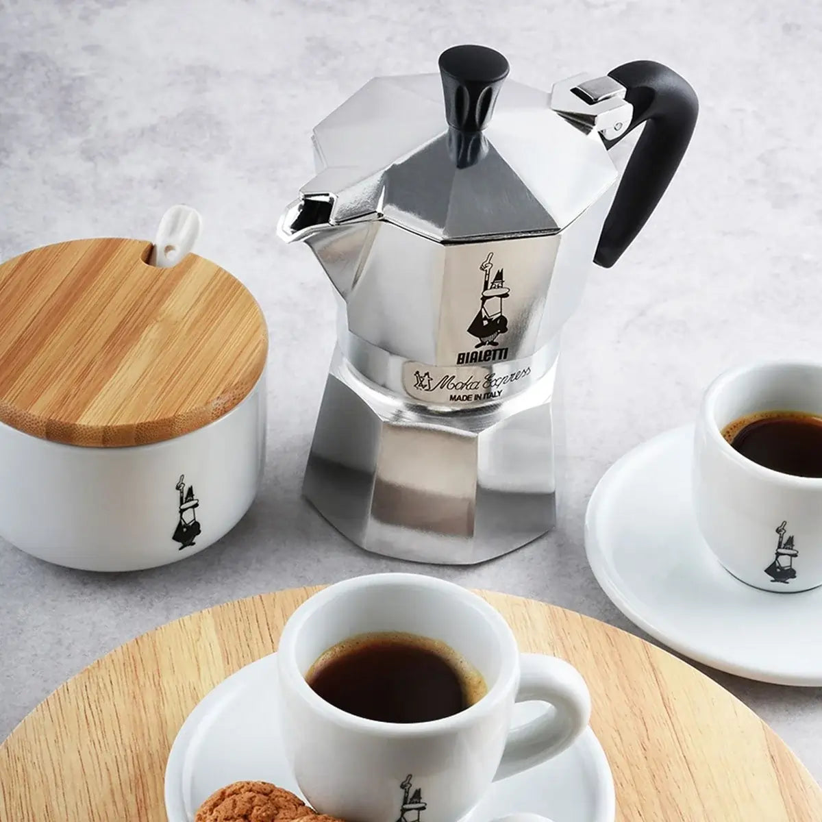  Kawiarka Bialetti Moka Express z filiżankami espresso i cukiernicą – włoski zestaw do parzenia kawy na tle jasnego blatu.