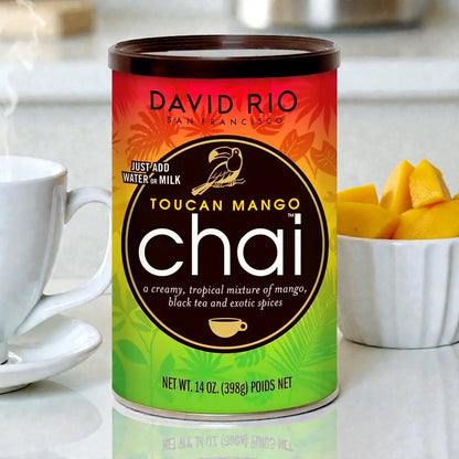 David Rio Toucan Mango Chai 398 g w ujęciu lifestyle – puszka chai o smaku mango ustawiona w kuchni obok filiżanki i kawałków świeżego mango