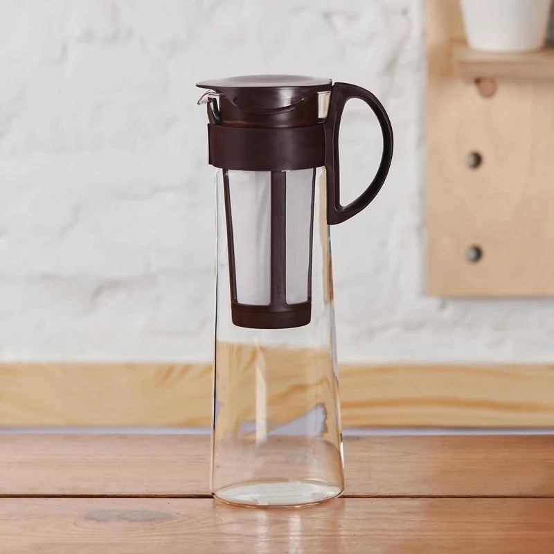  Dzbanek Hario do cold brew o pojemności 1 litra ustawiony na drewnianym stole – minimalistyczny design z brązową pokrywką i filtrem, idealny do domowego parzenia kawy na zimno
