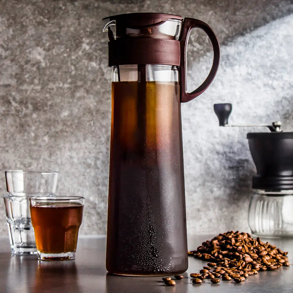 Dzbanek Hario Mizudashi z cold brew coffee – zaparzana na zimno kawa w eleganckim szklanym naczyniu z bordową pokrywką, obok szklanki i ziaren kawy