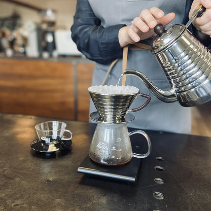 Parzenie kawy metodą przelewową: czajnik z wąską wylewką nalewa wodę do drippera Kalita Wave z papierowym filtrem na szklanym serwerze ustawionym na wadze.