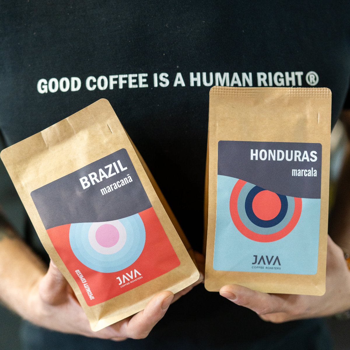 Osoba trzymająca dwie paczki kawy JAVA Coffee Roasters – Brazil Maracanã i Honduras Marcala. Na koszulce napis „Good coffee is a human right”