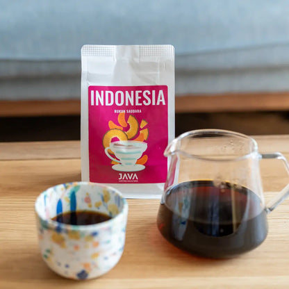 Kawa Indonesia Rukun Saudara od JAVA Coffee Roasters – opakowanie, kolorowa filiżanka i szklany serwer z zaparzoną kawą na stole w domowym wnętrzu