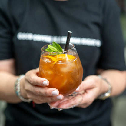 Orzeźwiająca cascara na lodzie z plastrem pomarańczy i miętą – letni napój kawowy w szklance, trzymany w dłoniach od JAVA Coffee Roasters
