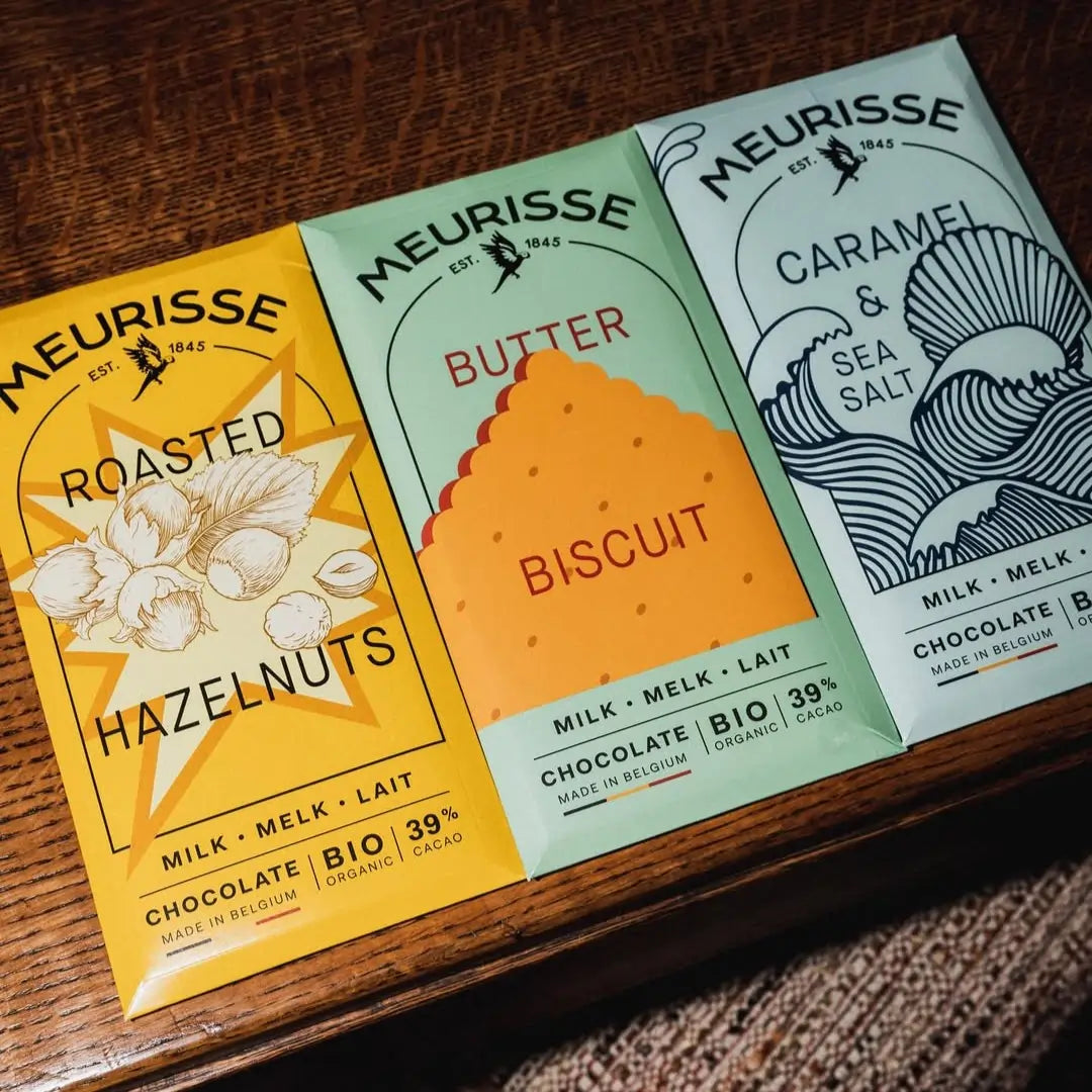 Trzy tabliczki mlecznej czekolady Meurisse: Roasted Hazelnuts, Butter Biscuit i Caramel & Sea Salt, ułożone na drewnianym stole w kolorowych opakowaniach z oznaczeniem BIO i Fairtrade.
