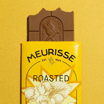 Tabliczka mlecznej czekolady Meurisse Roasted Hazelnuts 39% kakao częściowo wysunięta z żółtego opakowania z ilustracją orzechów laskowych, na tle w odcieniu żółtym.