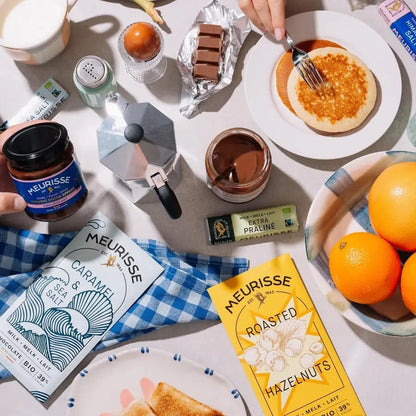 Śniadaniowa kompozycja z tabliczką mlecznej czekolady Meurisse Roasted Hazelnuts 39% kakao, Caramel & Sea Salt, kawą, naleśnikami i owocami na jasnym tle.