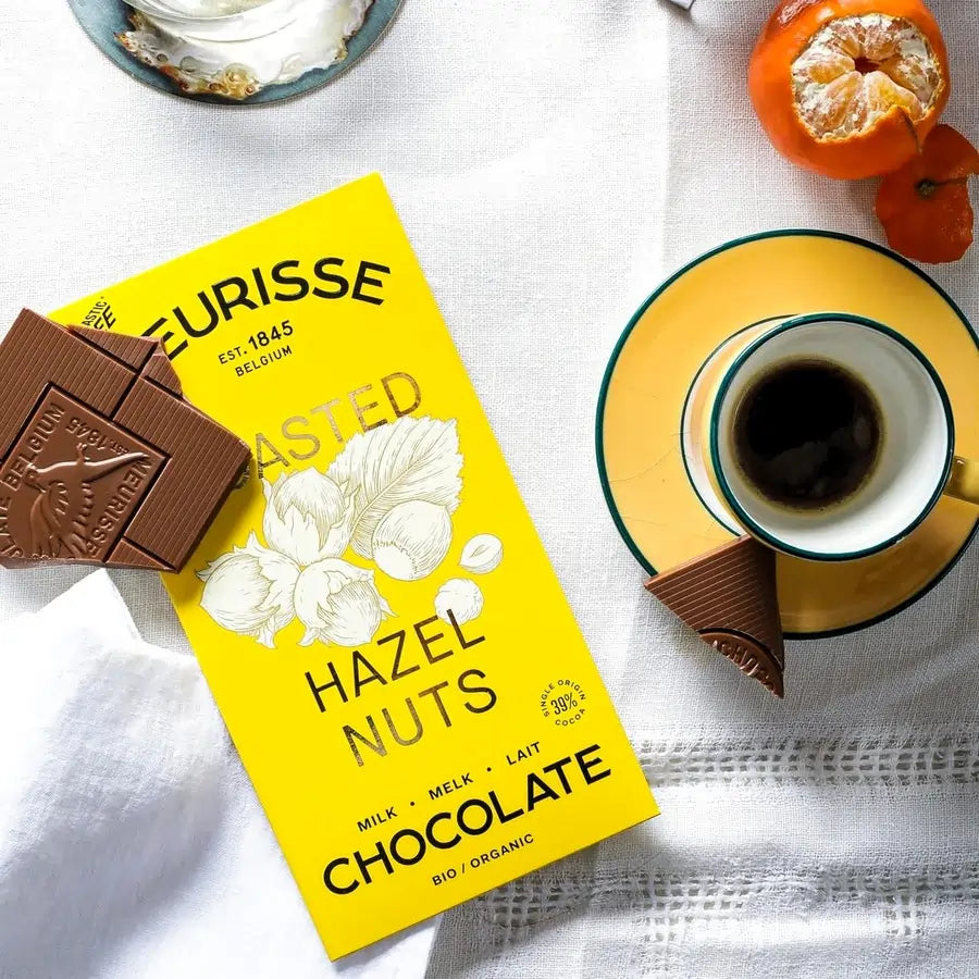 Tabliczka mlecznej czekolady Meurisse Roasted Hazelnuts 39% kakao w żółtym opakowaniu obok filiżanki kawy i kawałków czekolady na białym obrusie.