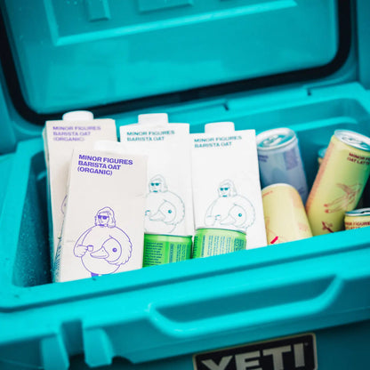 Kartony napoju owsianego Minor Figures Barista Oat i Barista Oat Organic w turkusowej lodówce turystycznej YETI – obok puszki z gotowymi napojami kawowymi na bazie owsa.