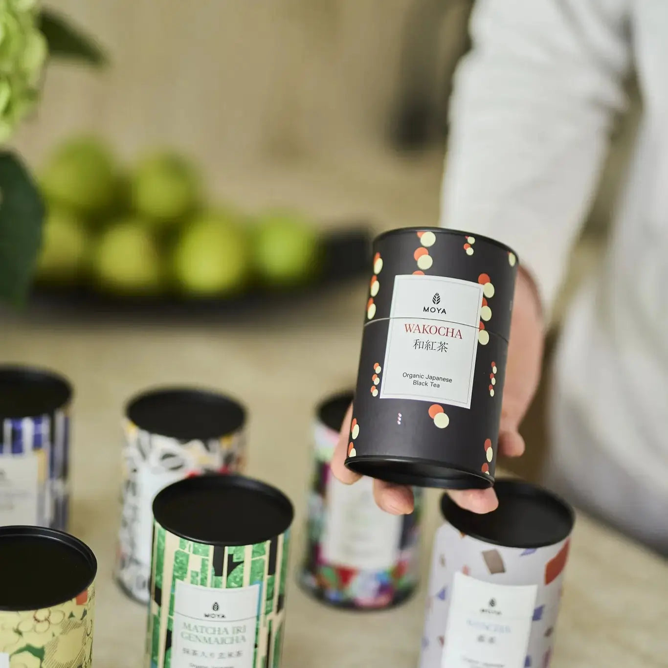 MOYA Wakocha – organiczna japońska czarna herbata trzymana w ręku, w otoczeniu innych herbat liściastych z kolekcji MOYA