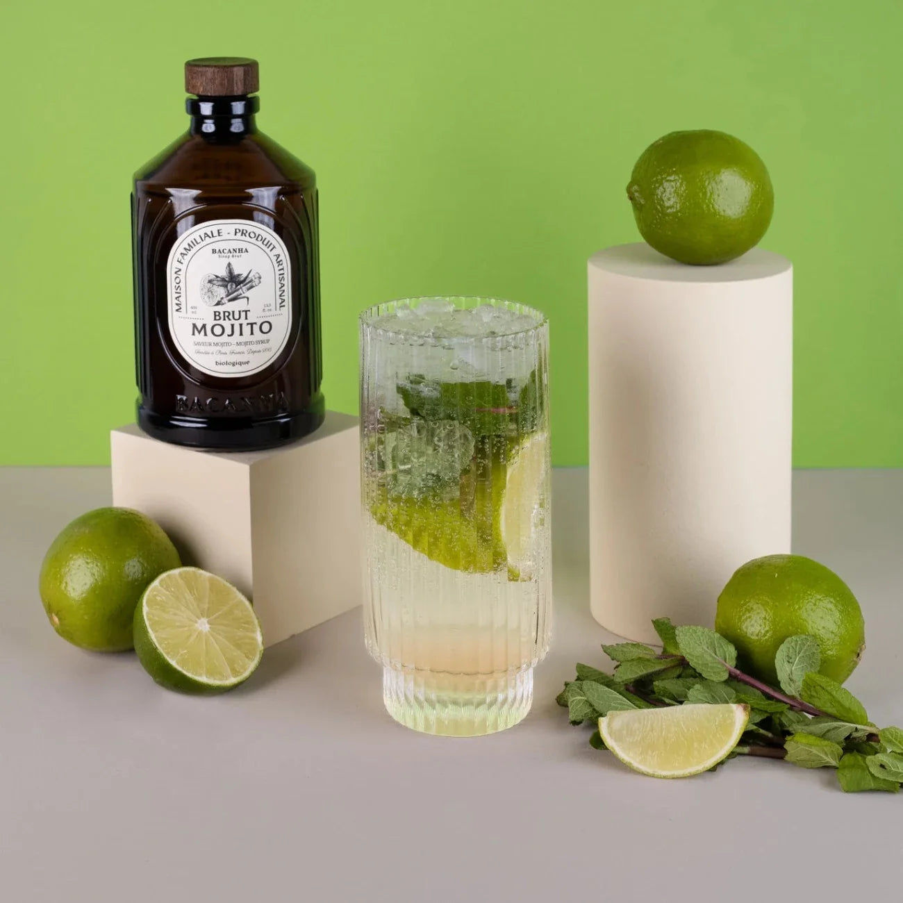 Syrop Bacanha Brut Mojito w butelce obok szklanki mojito z limonką, miętą i lodem – minimalistyczna aranżacja na zielonym tle.