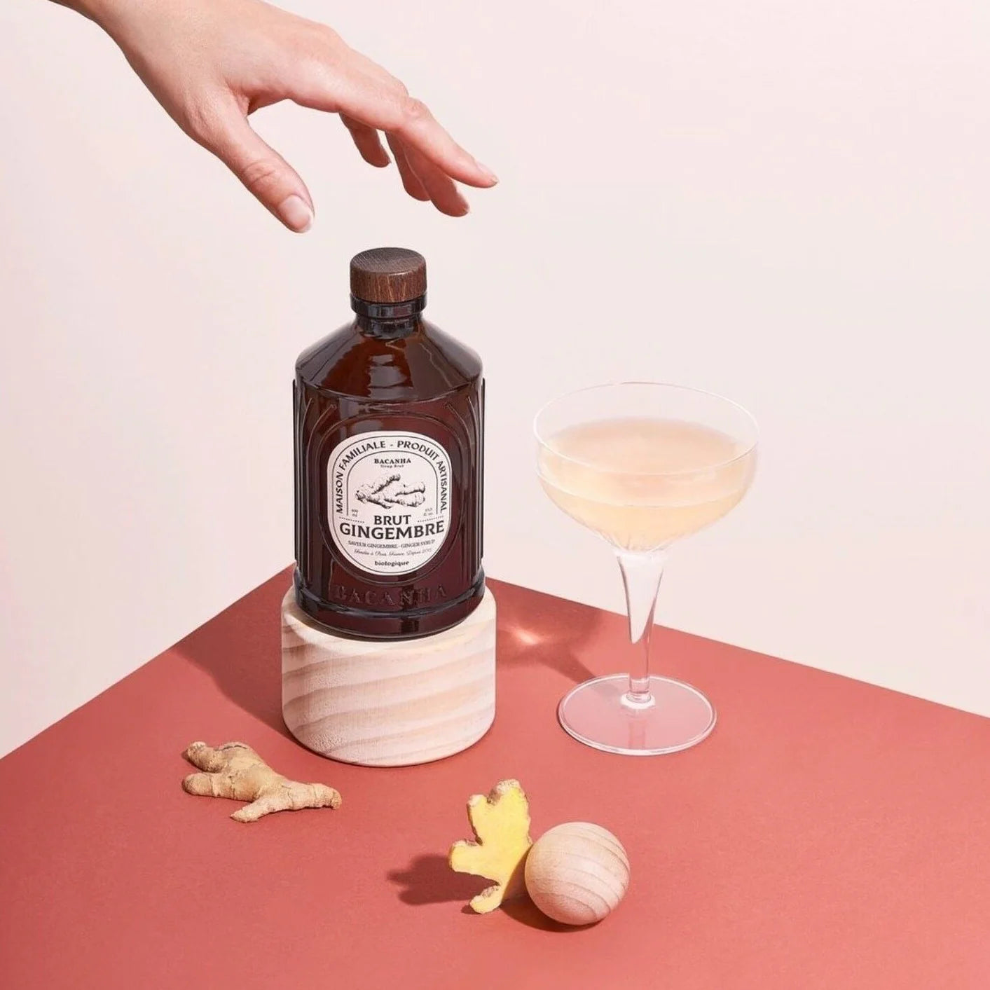 Syrop Bacanha Brut Gingembre (Imbirowy) w szklanej butelce, obok kieliszka z napojem i kawałków świeżego imbiru – elegancka kompozycja na tle w odcieniach różu.