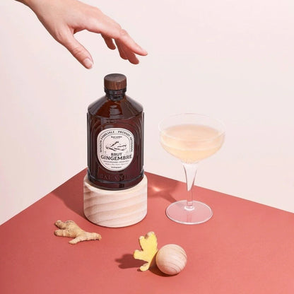 Syrop Bacanha Brut Gingembre (Imbirowy) w szklanej butelce, obok kieliszka z napojem i kawałków świeżego imbiru – elegancka kompozycja na tle w odcieniach różu.
