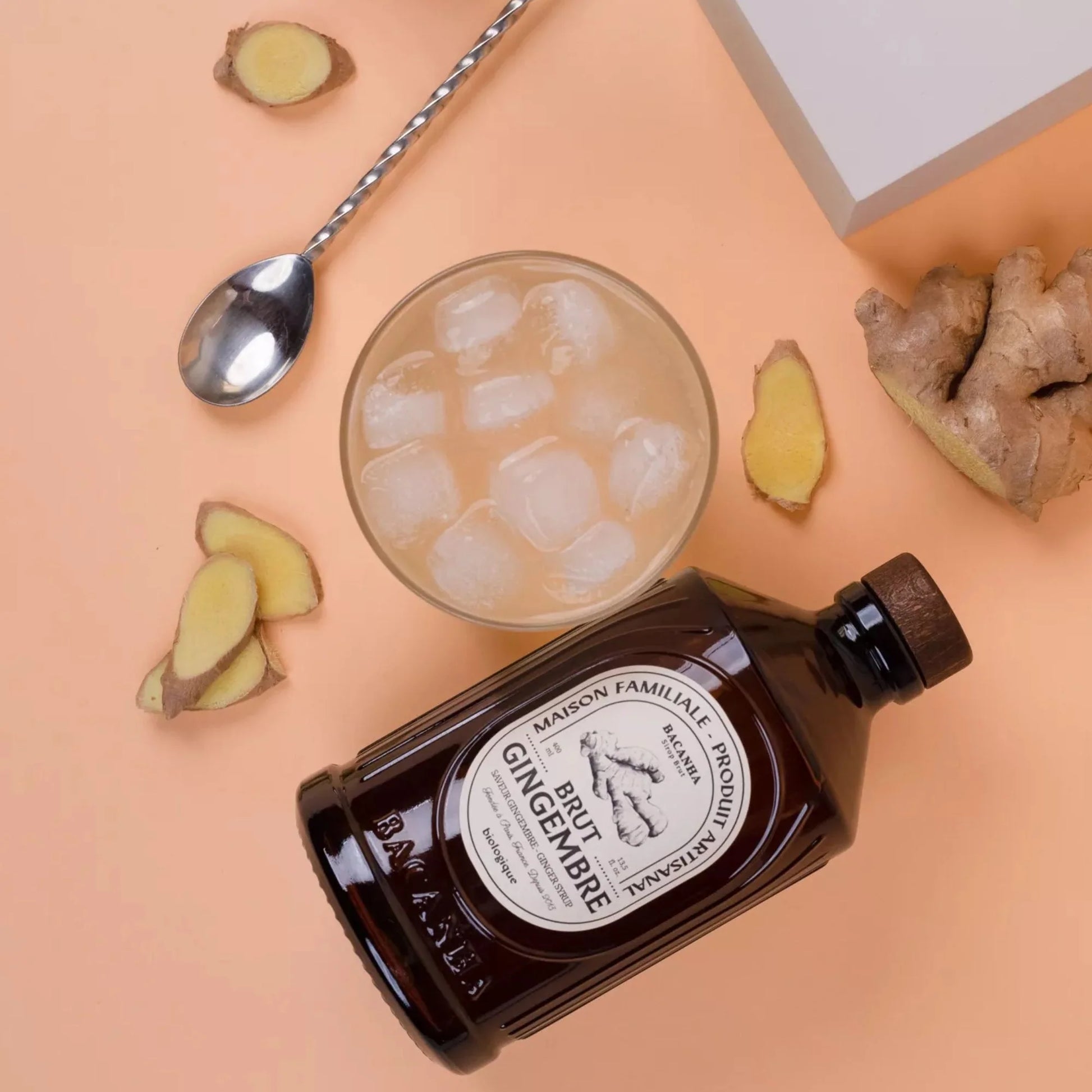 Syrop Bacanha Brut Gingembre (Imbirowy) z lodowym napojem i plasterkami świeżego imbiru – stylowa aranżacja na brzoskwiniowym tle.