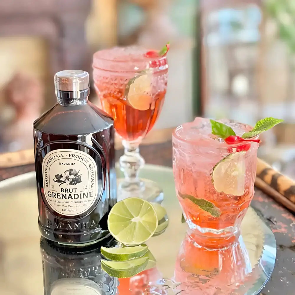 Syrop Grenadine Bacanha w szklanej butelce na tacy z lodowym koktajlem z limonką i wiśnią – orzeźwiający napój z dodatkiem organicznego syropu o smaku czerwonych owoców.