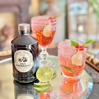 Syrop Grenadine Bacanha w szklanej butelce na tacy z lodowym koktajlem z limonką i wiśnią – orzeźwiający napój z dodatkiem organicznego syropu o smaku czerwonych owoców.