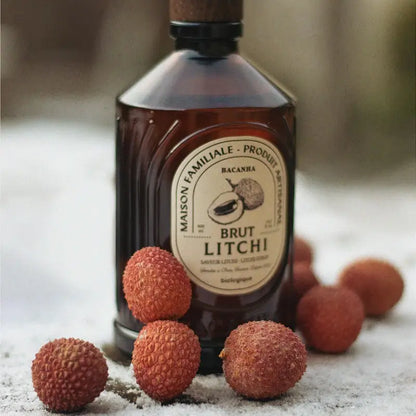 Syrop organiczny Bacanha o smaku liczi – butelka syropu na tle świeżych owoców liczi, ustawiona na śniegu.